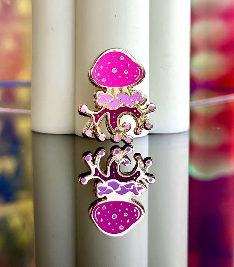 Op de afbeelding: Een gouden emaille pin met een roze en paarse paddenstoel met een draaiende steel. De paddenstoel heeft witte stippen en een gouden omtrek.