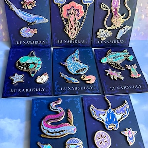 Op de afbeelding: Set van 8 emaillepins met verschillende zeedieren met een galaxy-achtig ontwerp. De pins zijn op blauwe kaartjes met gouden rand en de tekst "LUNARJELLY" gedrukt op elk kaartje.
