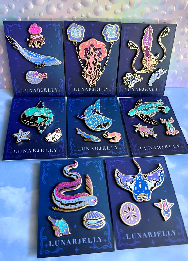 Op de afbeelding: Set van 8 emaillepins met verschillende zeedieren met een galaxy-achtig ontwerp. De pins zijn op blauwe kaartjes met gouden rand en de tekst "LUNARJELLY" gedrukt op elk kaartje.