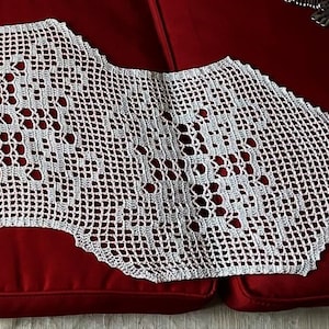 May include: White crocheted lace table runner with a scalloped edge and a repeating diamond pattern. The runner is laid out on a red surface.