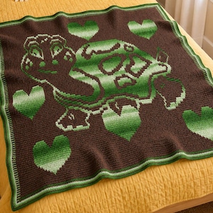 Op de afbeelding: Een vierkante gehaakte deken met een donkerbruine achtergrond en een centraal groen schildpadmotief. Het schild en het lichaam van de schildpad zijn uitgevoerd in diverse groentinten. Vijf groene ombre harten zijn rondom de schildpad geplaatst. De deken heeft een groene rand.