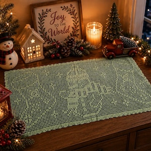 May include: A sage green, rectangular, crocheted table runner with a detailed snowflake design. The runner is displayed on a wooden surface, surrounded by Christmas decorations, including a snowman, a miniature red truck, and a sign that reads "Joy to the World."