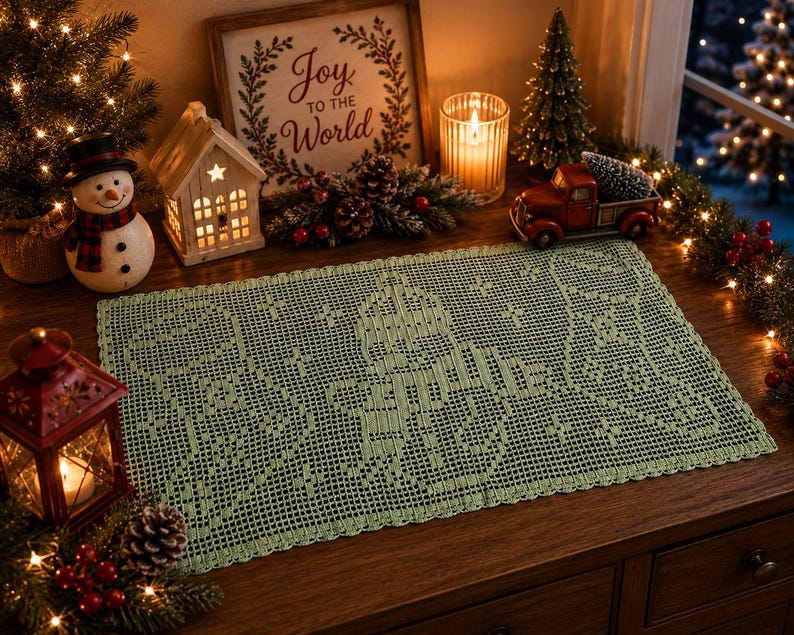May include: A sage green, rectangular, crocheted table runner with a detailed snowflake design. The runner is displayed on a wooden surface, surrounded by Christmas decorations, including a snowman, a miniature red truck, and a sign that reads "Joy to the World."