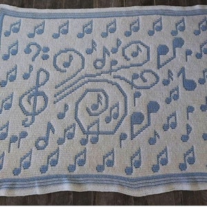 Puede incluir: Una manta de crochet blanca con un patrón de notas musicales azules. La manta presenta una variedad de notas musicales, incluyendo claves de sol y corcheas.