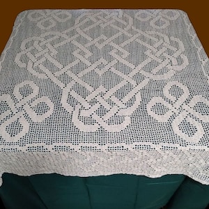 Puede incluir: Un mantel de mesa blanco de ganchillo con un delicado patrón floral. El mantel está colocado sobre una superficie verde oscuro.