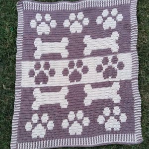 Può includere: Una coperta rettangolare all'uncinetto con un motivo ripetuto di impronte di zampe bianche e ossi di cane bianchi su sfondo color malva. La coperta è rifinita con un bordo a righe in punti alternati color malva e bianco su tutti i lati.