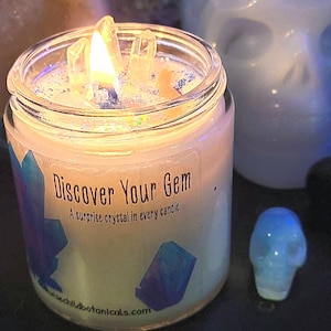 May include: A clear glass candle jar with a white candle and a crystal inside. The candle is lit and the jar has the text "Discover Your Gem: A surprise crystal in every candle" on the label. The candle is sitting on a black surface next to a blue crystal skull.