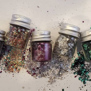 Puede incluir: Cinco pequeños frascos de vidrio llenos de purpurina de diferentes colores. Los frascos tienen tapas plateadas y están dispuestos en fila. La purpurina está esparcida alrededor de los frascos.