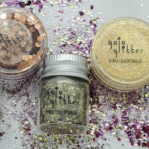 Puede incluir: Cuatro pequeños frascos de vidrio llenos de purpurina de diferentes colores. Los frascos están etiquetados con "Gaia Glitter by Wild Child Botanicals".