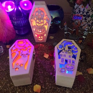 Peut inclure: Trois bo&icirc;tes en bois blanc en forme de cercueil avec des motifs d'Halloween lumineux. La premi&egrave;re bo&icirc;te a une pile de citrouilles, la deuxi&egrave;me a un chat noir avec des &eacute;toiles et la troisi&egrave;me a une maison hant&eacute;e avec des citrouilles.