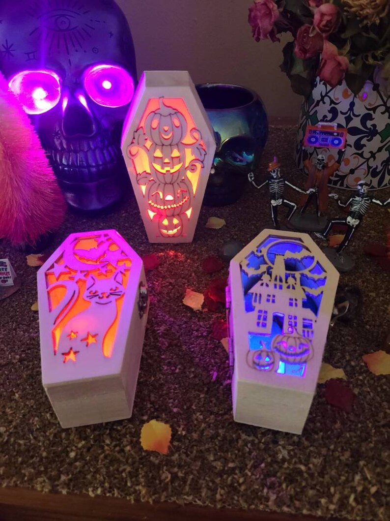 Peut inclure: Trois bo&icirc;tes en bois blanc en forme de cercueil avec des motifs d'Halloween lumineux. La premi&egrave;re bo&icirc;te a une pile de citrouilles, la deuxi&egrave;me a un chat noir avec des &eacute;toiles et la troisi&egrave;me a une maison hant&eacute;e avec des citrouilles.