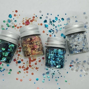 May include: Four small glass jars filled with different colored glitter. The jars have silver lids and are arranged in a row on a white surface. The glitter colors are teal, orange, blue, and white.