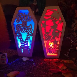 Peut inclure: Deux d&eacute;corations d'Halloween en bois en forme de cercueil avec des fonctions lumineuses. Le cercueil de gauche est bleu et dit "Trick or Treat" avec des jambes et un chapeau de sorci&egrave;re. Le cercueil de droite est rouge et dit "Hocus Pocus".