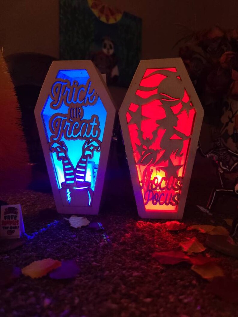 Peut inclure: Deux d&eacute;corations d'Halloween en bois en forme de cercueil avec des fonctions lumineuses. Le cercueil de gauche est bleu et dit "Trick or Treat" avec des jambes et un chapeau de sorci&egrave;re. Le cercueil de droite est rouge et dit "Hocus Pocus".