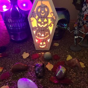 Peut inclure: Une bo&icirc;te en bois en forme de cercueil avec une lumi&egrave;re &agrave; l'int&eacute;rieur, pr&eacute;sentant une pile de visages de citrouilles sculpt&eacute;s. La bo&icirc;te est pos&eacute;e sur une surface recouverte de petites pierres et feuilles color&eacute;es.