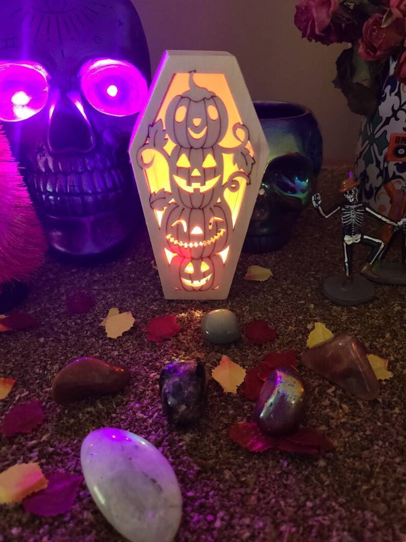 Peut inclure: Une bo&icirc;te en bois en forme de cercueil avec une lumi&egrave;re &agrave; l'int&eacute;rieur, pr&eacute;sentant une pile de visages de citrouilles sculpt&eacute;s. La bo&icirc;te est pos&eacute;e sur une surface recouverte de petites pierres et feuilles color&eacute;es.