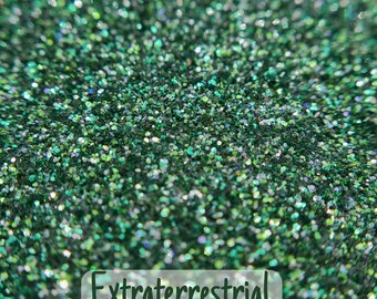 May include: Close-up of green glitter with iridescent flecks. The text "Extraterrestrial" is displayed in a white font at the bottom of the image. The glitter is scattered and appears to be a fine grain.