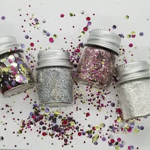 Puede incluir: Cuatro pequeños frascos de vidrio llenos de purpurina de diferentes colores. Los frascos tienen tapas plateadas y están dispuestos sobre una superficie blanca con purpurina esparcida.