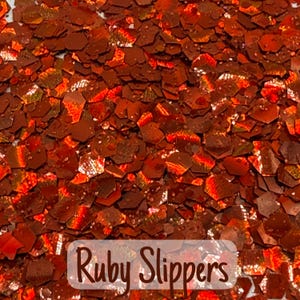 May include: Close-up of a pile of ruby red glitter flakes. The flakes are hexagonal and reflect light, creating a sparkling effect. The text "Ruby Slippers" is displayed at the bottom of the image.