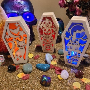 Peut inclure: Trois cercueils lumineux en bois sur le th&egrave;me d'Halloween. Les cercueils sont d&eacute;cor&eacute;s d'un chat, de citrouilles et d'une maison hant&eacute;e. Les cercueils sont &eacute;clair&eacute;s par des lumi&egrave;res orange, rouge et bleue.