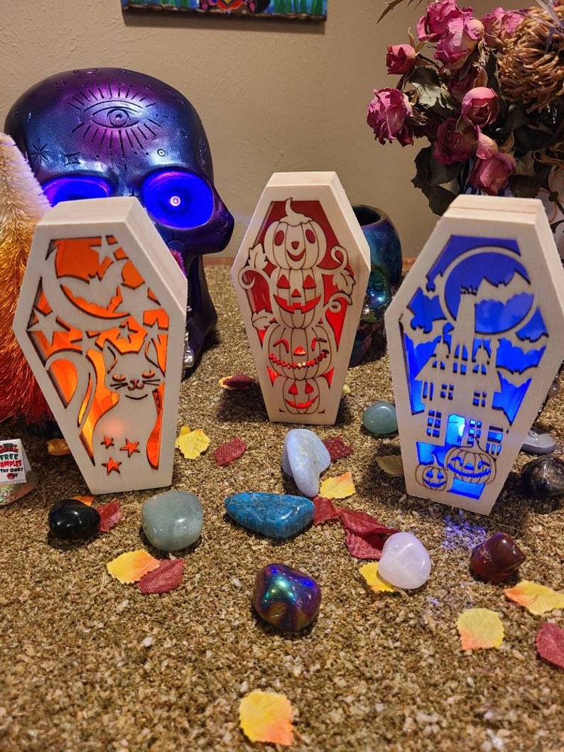 Peut inclure: Trois cercueils lumineux en bois sur le th&egrave;me d'Halloween. Les cercueils sont d&eacute;cor&eacute;s d'un chat, de citrouilles et d'une maison hant&eacute;e. Les cercueils sont &eacute;clair&eacute;s par des lumi&egrave;res orange, rouge et bleue.