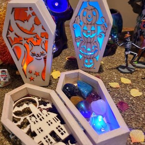 Peut inclure: Trois bo&icirc;tes en bois en forme de cercueil avec des motifs sur le th&egrave;me d'Halloween. La premi&egrave;re bo&icirc;te a un chat et une lune croissante avec des &eacute;toiles. La deuxi&egrave;me bo&icirc;te a une pile de trois citrouilles. La troisi&egrave;me bo&icirc;te a une maison hant&eacute;e et une citrouille. La troisi&egrave;me bo&icirc;te est ouverte et contient une vari&eacute;t&eacute; de pierres pr&eacute;cieuses color&eacute;es.