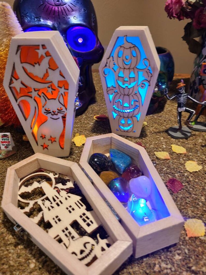 Peut inclure: Trois bo&icirc;tes en bois en forme de cercueil avec des motifs sur le th&egrave;me d'Halloween. La premi&egrave;re bo&icirc;te a un chat et une lune croissante avec des &eacute;toiles. La deuxi&egrave;me bo&icirc;te a une pile de trois citrouilles. La troisi&egrave;me bo&icirc;te a une maison hant&eacute;e et une citrouille. La troisi&egrave;me bo&icirc;te est ouverte et contient une vari&eacute;t&eacute; de pierres pr&eacute;cieuses color&eacute;es.