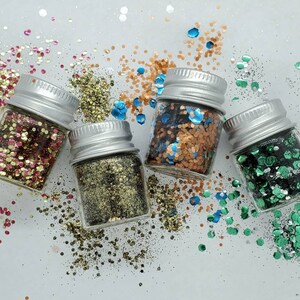May include: Five small glass jars filled with different colored glitter. The jars have silver lids and are arranged in a semi-circle on a white surface. The glitter colors are gold, green, red, blue, and copper.