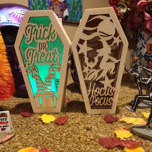 Peut inclure: Deux cercueils lumineux en bois sur le th&egrave;me d'Halloween. Un cercueil est vert et dit "Trick or Treat" avec des jambes de sorci&egrave;re et un chaudron. L'autre cercueil est brun et dit "Hocus Pocus" avec une silhouette de sorci&egrave;re.