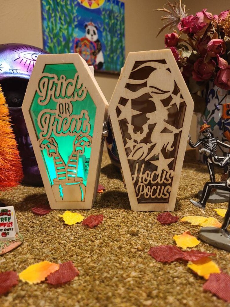 Peut inclure: Deux cercueils lumineux en bois sur le th&egrave;me d'Halloween. Un cercueil est vert et dit "Trick or Treat" avec des jambes de sorci&egrave;re et un chaudron. L'autre cercueil est brun et dit "Hocus Pocus" avec une silhouette de sorci&egrave;re.