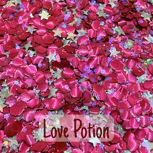 Puede incluir: Primer plano de una mezcla de purpurina rosa y roja, que incluye piezas en forma de estrella y pétalo. El texto "Love Potion" es visible en fuente blanca sobre la purpurina. La purpurina tiene una calidad brillante e iridiscente.