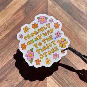 May include: A die-cut holographic sticker with a white background featuring orange and pink flowers and the text "PROBABLY ON MY WAY TO THE THRIFT STORE" in yellow letters with a dark outline. The sticker has a scalloped edge and a glittery finish.