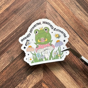 May include: A sticker featuring a green frog wearing a crown, sitting on a pink mushroom, surrounded by flowers and insects. The text reads "Naturally Introverted, Selectively Extroverted."