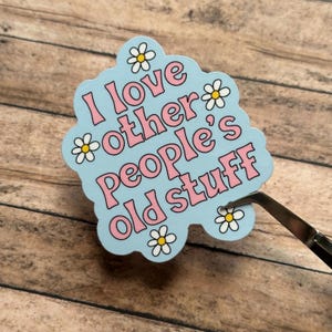 May include: A light blue die-cut sticker featuring the pink text I love other people's old stuff. The sticker has a scalloped edge and is decorated with several small white daisies with yellow centers. A pair of metal tweezers holds the sticker.