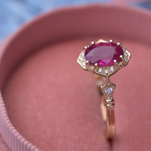 Puede incluir: Un anillo de oro con una gran piedra de rubí ovalada rodeada por un halo de diamantes más pequeños. El anillo está engastado en un estilo vintage con una banda delicada.