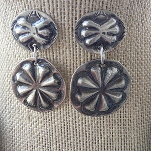 May include: A pair of silver-toned drop earrings featuring two circular discs each. The top disc has a four-petaled embossed design, while the larger bottom disc displays an eight-petaled floral pattern. Both discs have decorative stamped edges.