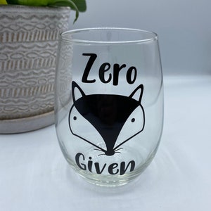 以下が含まれることがあります： 黒いキツネのデザインと「Zero Given」という文字が印刷された透明なガラス製のワイングラスです。