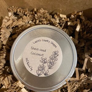 May include: A silver tin with a brown label that reads "Laurel's Country Shoppe Shea and Coconut 100% Natural Soy". The label has a drawing of a plant with leaves.