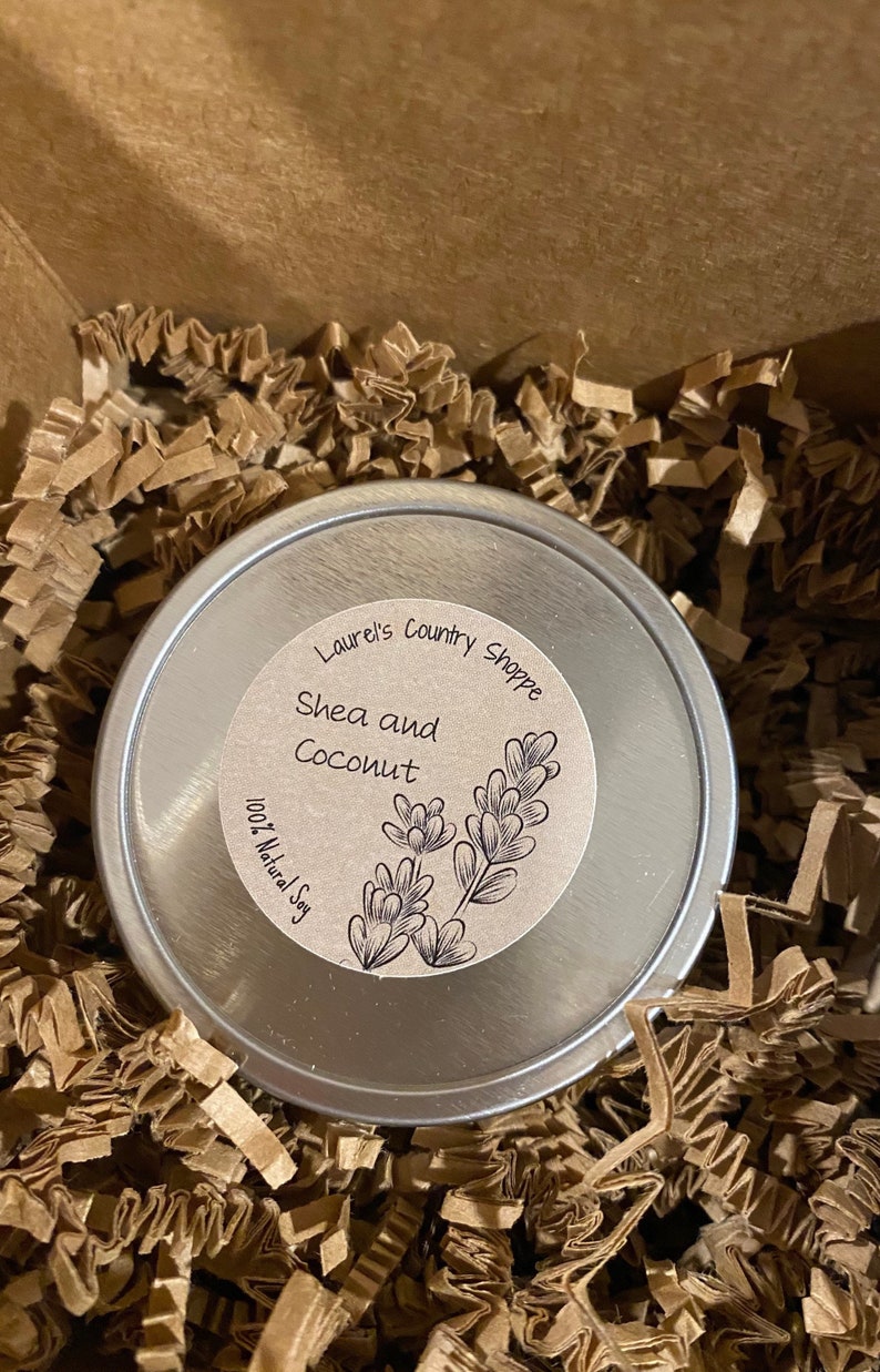 May include: A silver tin with a brown label that reads "Laurel's Country Shoppe Shea and Coconut 100% Natural Soy". The label has a drawing of a plant with leaves.