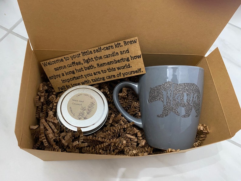 May include: A brown cardboard box filled with brown packing paper. The box contains a gray mug with a glitter bear design, a small tin of soap, and a card with the text "Welcome to your little self-care kit. Brew some coffee, light the candle and enjoy a long hot bath. Remembering how important you are to this world. Fall in love with taking care of yourself."
