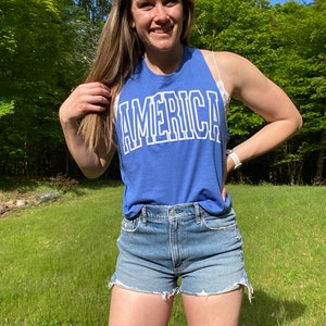 Puede incluir: Una mujer con una camiseta sin mangas azul con la palabra "AMERICA" impresa en blanco. Tambi&eacute;n lleva pantalones cortos de mezclilla.