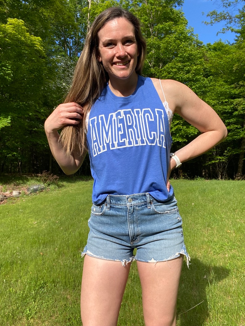 Puede incluir: Una mujer con una camiseta sin mangas azul con la palabra "AMERICA" impresa en blanco. Tambi&eacute;n lleva pantalones cortos de mezclilla.