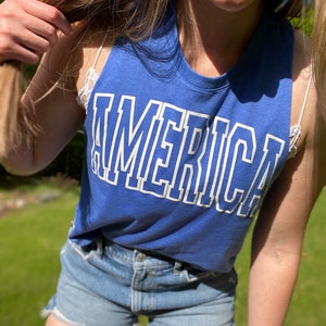 Puede incluir: Un top azul sin mangas con la palabra "AMERICA" impresa en blanco en la parte delantera. El top lo lleva una persona con el pelo largo rubio y pantalones cortos de mezclilla.