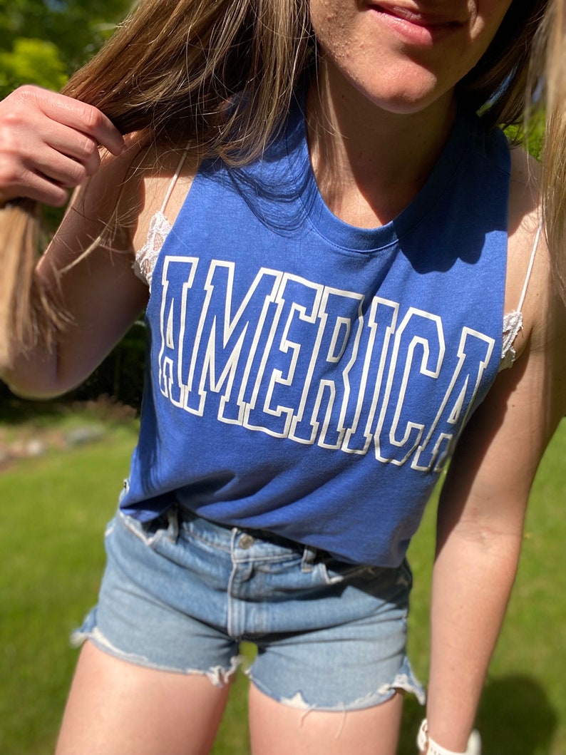 Puede incluir: Un top azul sin mangas con la palabra "AMERICA" impresa en blanco en la parte delantera. El top lo lleva una persona con el pelo largo rubio y pantalones cortos de mezclilla.