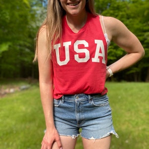 Puede incluir: Una mujer lleva una camiseta roja sin mangas con la palabra "USA" impresa en blanco en la parte delantera. Tambi&eacute;n lleva pantalones cortos de mezclilla.