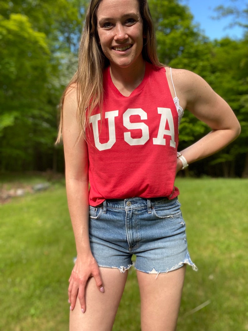 Puede incluir: Una mujer lleva una camiseta roja sin mangas con la palabra "USA" impresa en blanco en la parte delantera. Tambi&eacute;n lleva pantalones cortos de mezclilla.