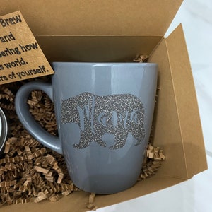 May include: A gray ceramic mug with a silver glitter bear and the word "Mama" on it. The mug is in a brown cardboard box with brown crinkle paper.