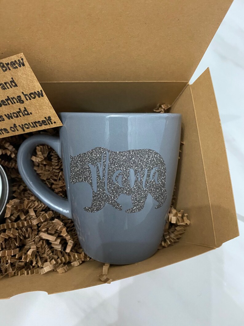 May include: A gray ceramic mug with a silver glitter bear and the word "Mama" on it. The mug is in a brown cardboard box with brown crinkle paper.