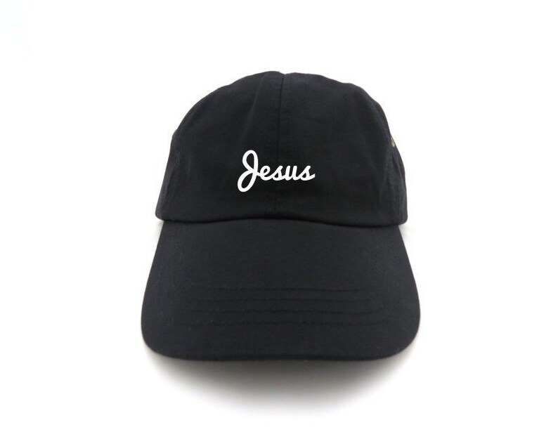 May include: Black baseball cap with a curved brim. The word "Jesus" is embroidered in white script on the front of the cap. The cap is made of a soft fabric and has an adjustable strap.