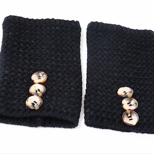 May include: Two black knitted wrist warmers with three round, tortoiseshell-colored buttons each. The wrist warmers are made of a textured, dark yarn and are displayed against a white background.